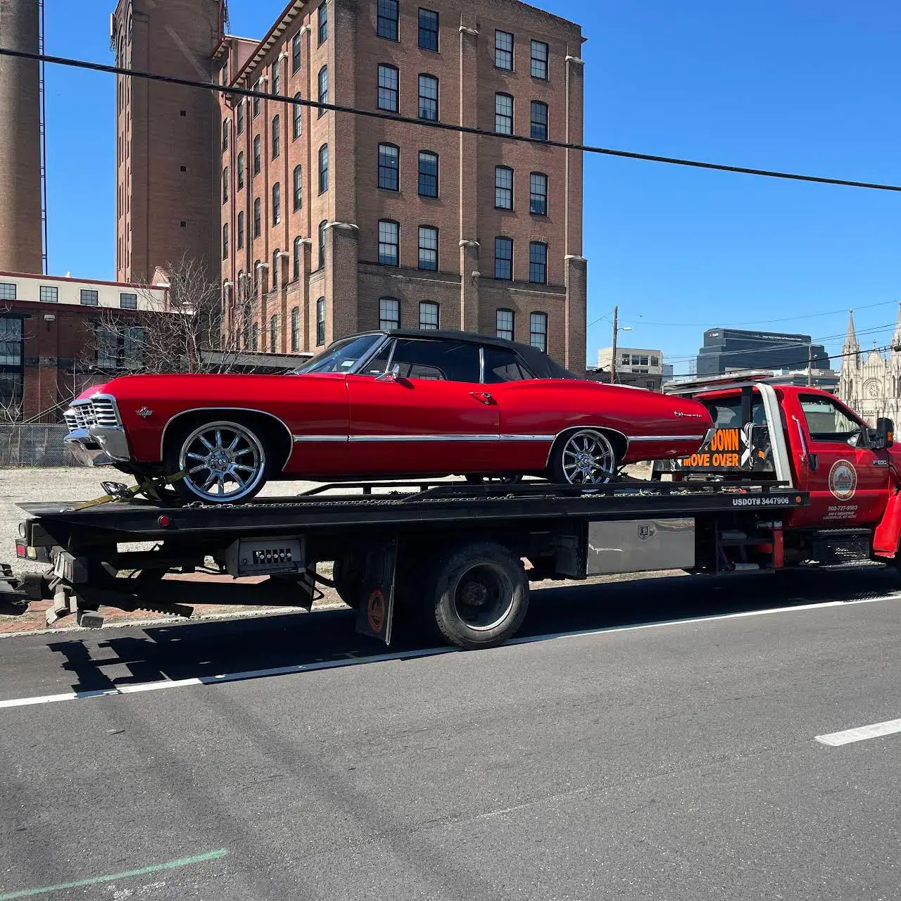 Leo's flatbed hauling a red classic convertible past a brick building